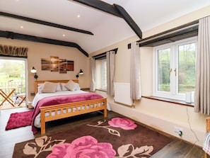A bedroom with a wooden bed, a large window with curtains, a radiator, and a view of greenery outside. - Cider Press Cottage (Torpoint)