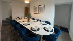 Uma área de refeições com uma mesa comprida, cadeiras azuis, pratos brancos e um vaso com folhagem.