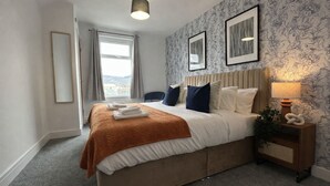 Quarto de hotel com cama, mesa de cabeceira, luminária e janela com cortinas.