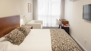 Habitación de hotel con cama, escritorio, silla, televisor y ventana con cortinas.