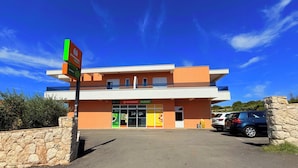 A two-story building with a sign for 'STUDENAC MARKET' and a parking area with cars. - Apartment Top 5 with sunny balcony and sea view (Novalja)
