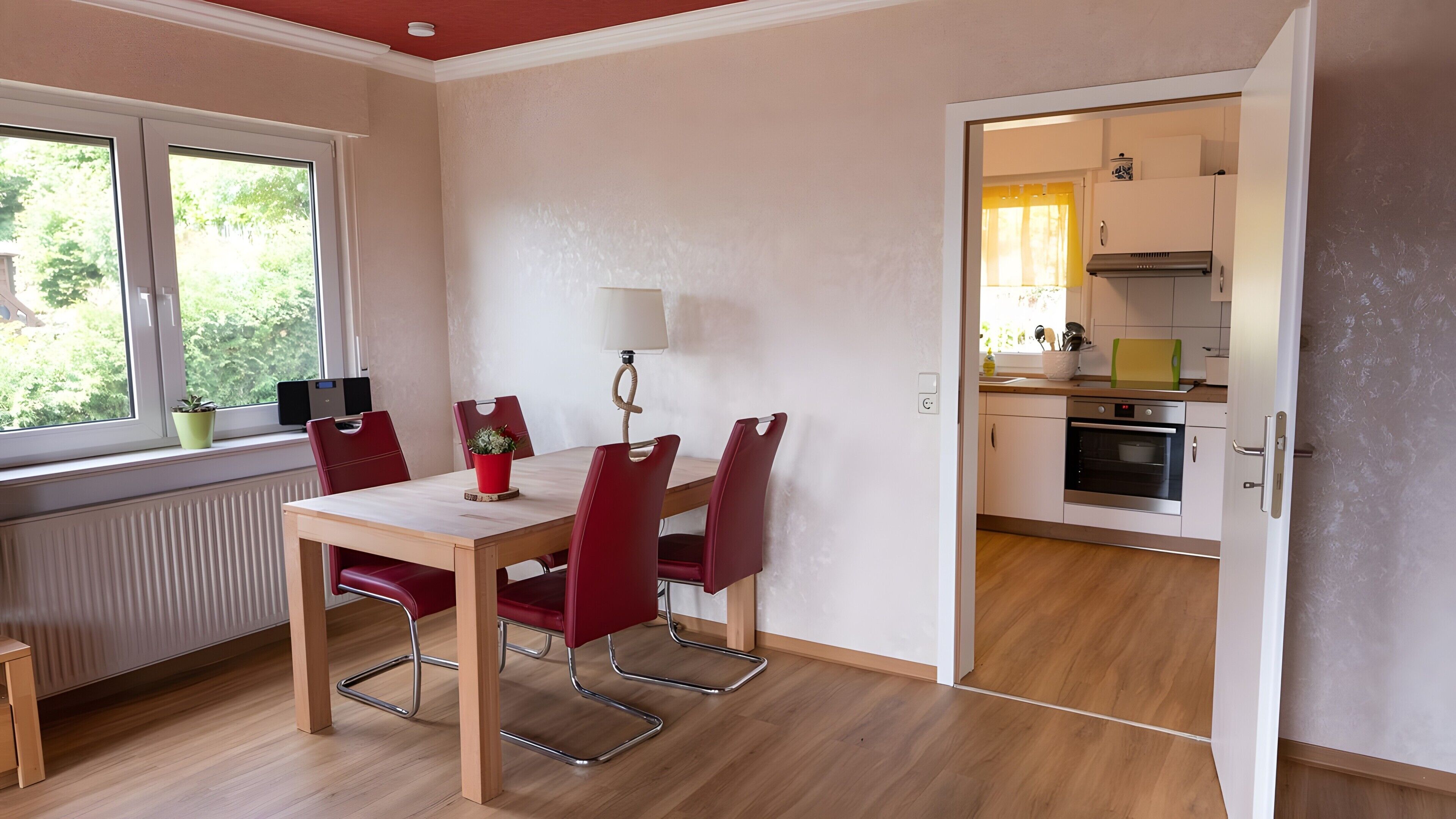 Un área de comedor con una mesa de madera y sillas rojas, una ventana con vistas a la vegetación, y una cocina con electrodomésticos modernos.