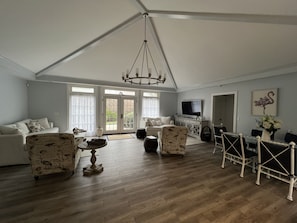 A spacious living room with a high ceiling, a chandelier, a television, a dining area with a table and chairs, and a large window with curtains.