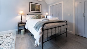 Une chambre à coucher comprenant un lit, deux tables de chevet, un climatiseur fixé au mur, un tableau encadré, une porte et un tapis à motifs.