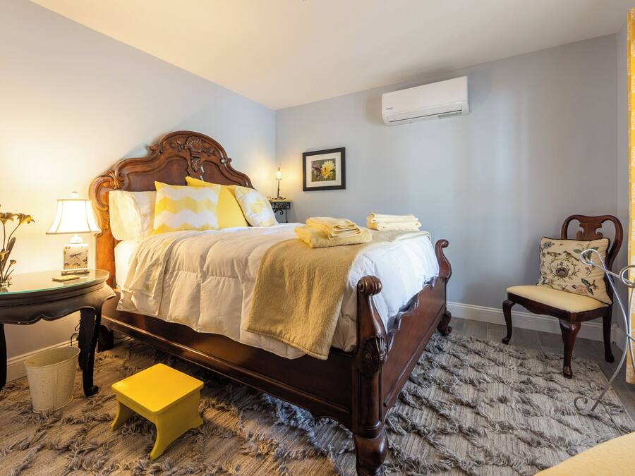 Un dormitorio con una cama de madera, una mesita de noche con lámpara, una silla, un taburete pequeño y un aire acondicionado instalado en la pared.