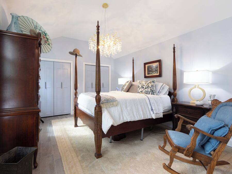 A bedroom with a four-poster bed, a wooden rocking chair, a dresser, and a chandelier.