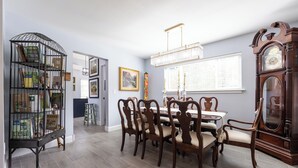 Une salle à manger avec une table en bois, six chaises, un lustre, une horloge comtoise et une bibliothèque.
