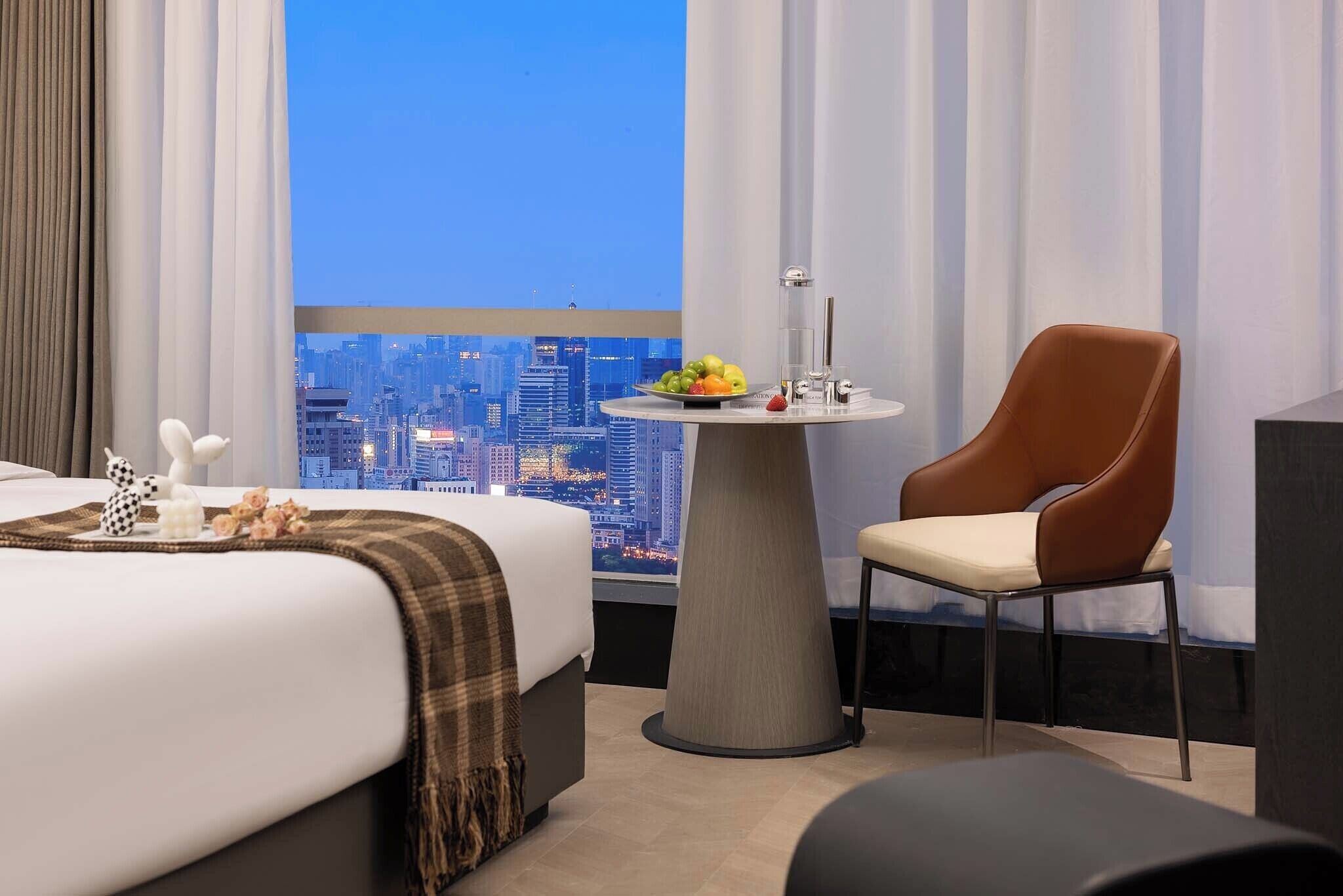 A hotel room with a bed, a round table with a fruit bowl, a brown leather chair, and a city view.