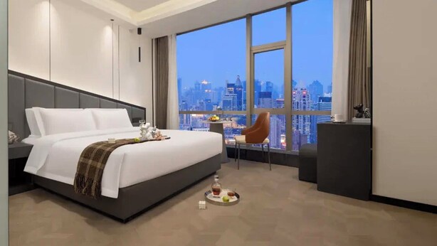 Een moderne hotelkamer met een groot bed, uitzicht op de stad, een bureau met stoel, en een kleine tafel met een fruitschaal.