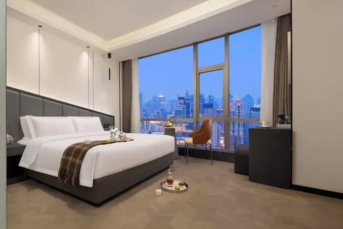 Ein modernes Hotelzimmer mit einem großen Bett, Blick auf die Stadt, einem Schreibtisch mit Stuhl und einem kleinen Tisch mit Obstteller.