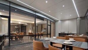 Een moderne eetruimte met glazen wanden, tafels gedekt met witte borden, en een keuken zichtbaar op de achtergrond.