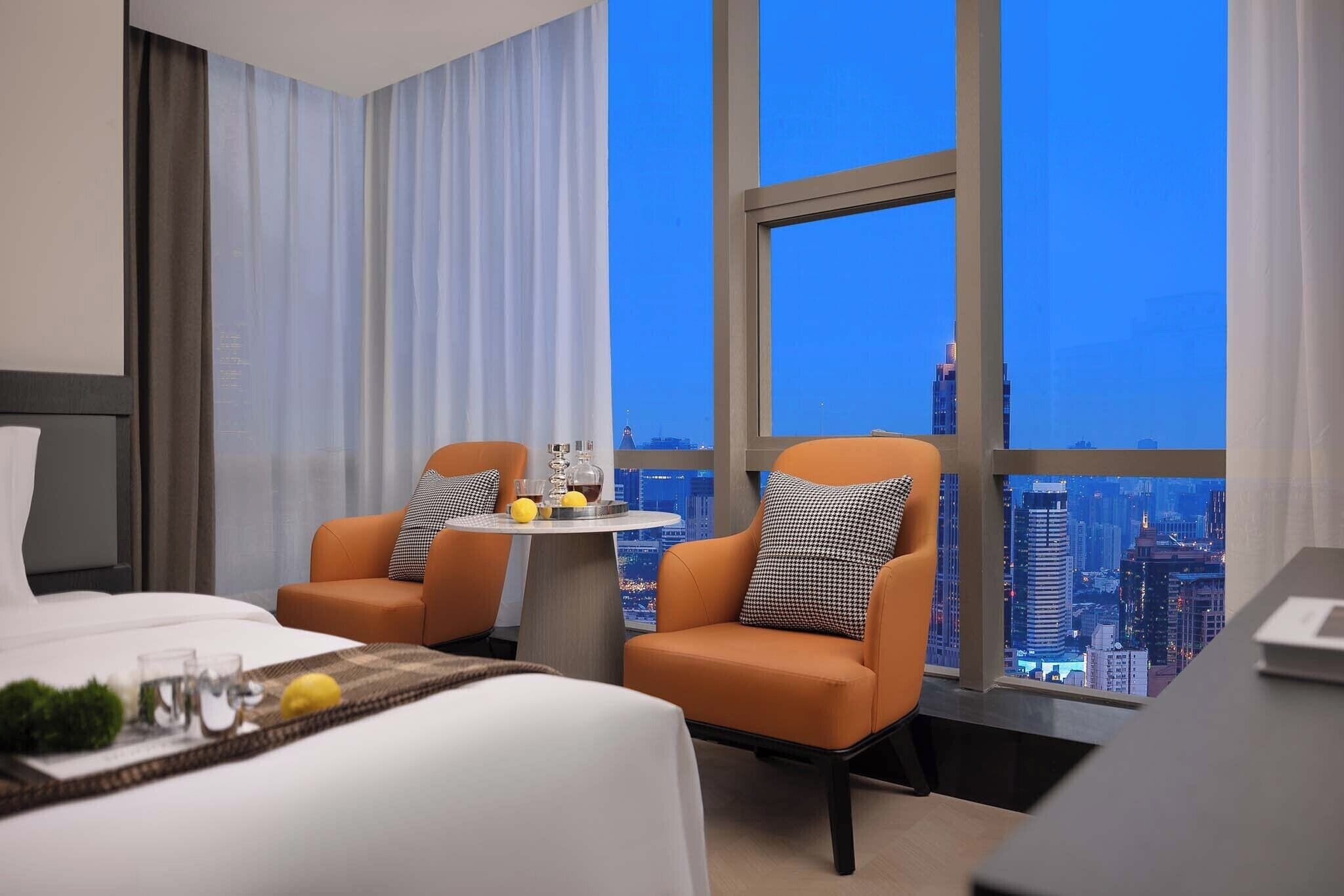A hotel room with a bed, two armchairs, a small table with drinks, and a city view through large windows.