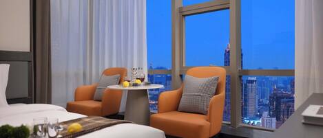 A hotel room with a bed, two armchairs, a small table with drinks, and a city view through large windows.