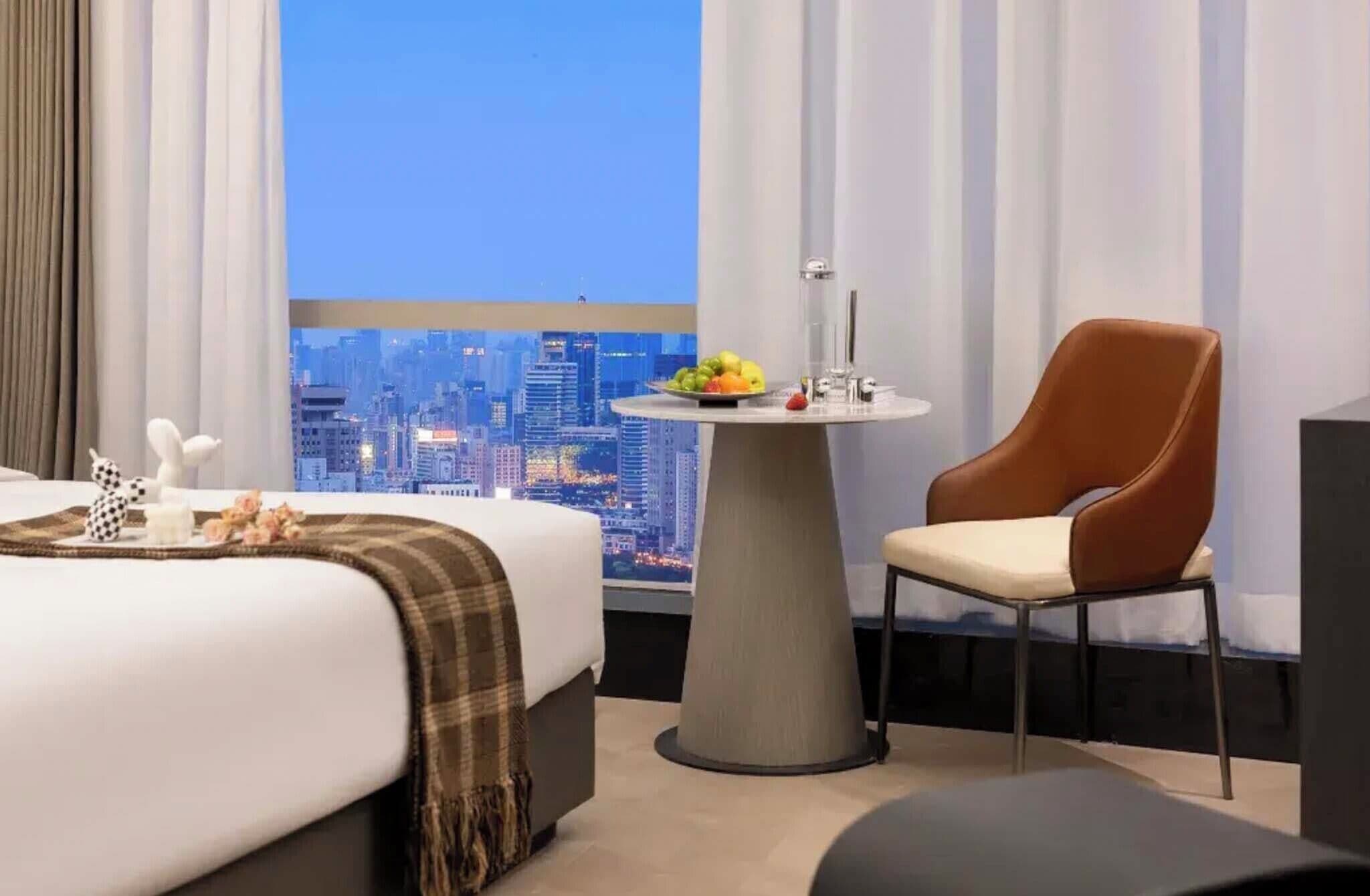 A hotel room with a bed, a round table with fruit, a chair, and a city view.