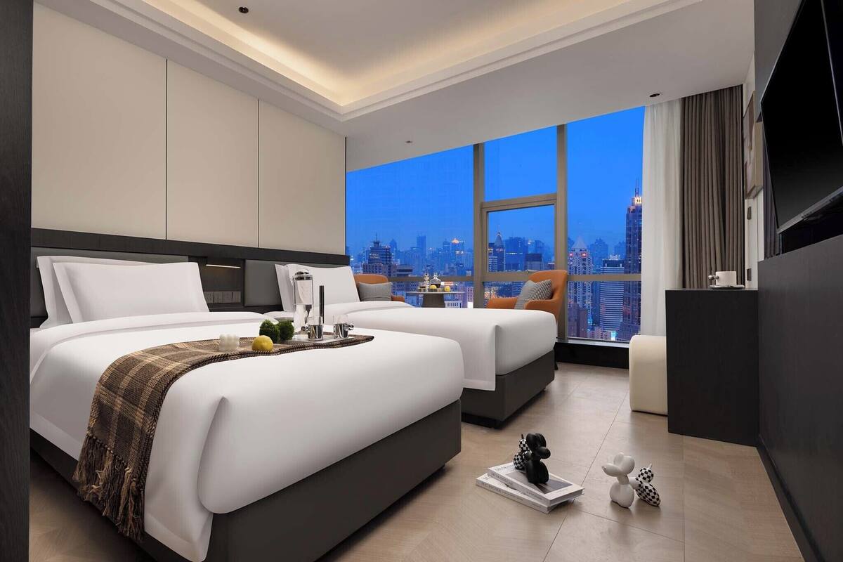 Ein modernes Hotelzimmer mit zwei Betten, einem großen Fenster mit Stadtblick, einem Flachbildfernseher und einem kleinen Tisch mit Dekoration.