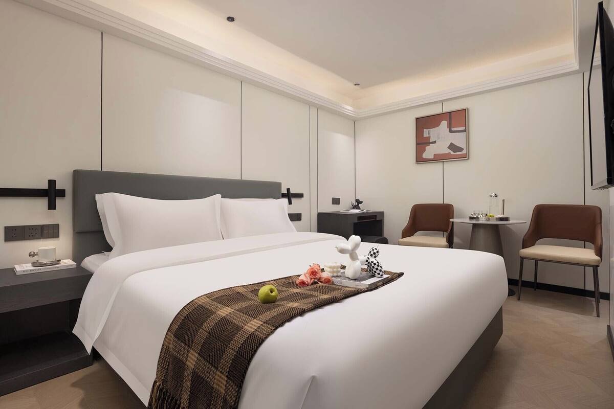 Ein Hotelzimmer mit einem großen Bett, einem Nachttisch, einem kleinen Schreibtisch, zwei Stühlen und einem an der Wand montierten Fernseher.