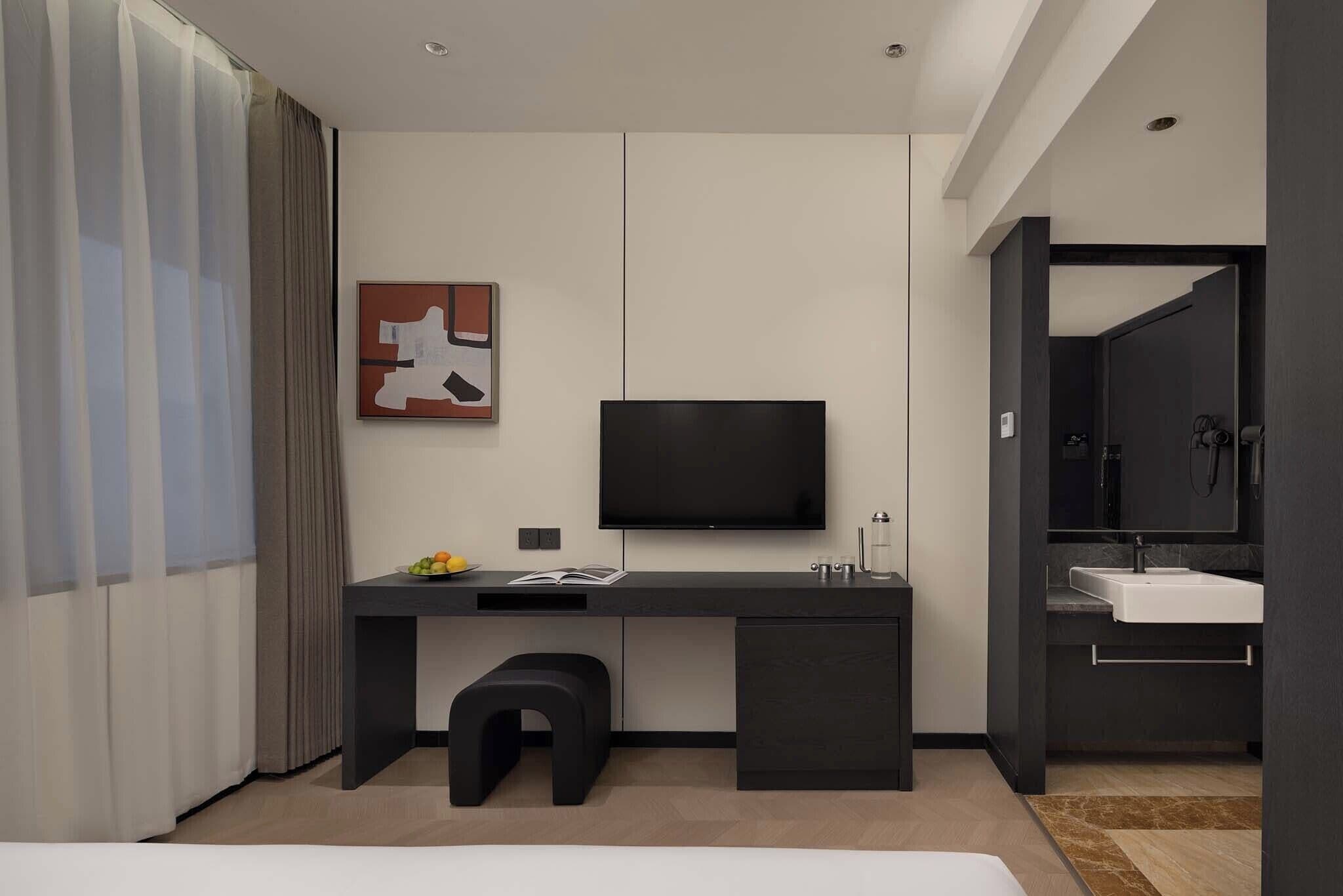 Une chambre d’hôtel moderne dotée d’un grand lit, d’un bureau avec un téléviseur à écran plat, d’un petit tabouret, d’une table d’appoint avec des fruits, et d’une salle de bain avec un lavabo et un miroir.