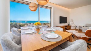 Een eethoek met een houten tafel gedekt voor vier personen, een Boeddhabeeld en een groot raam met uitzicht op zee.