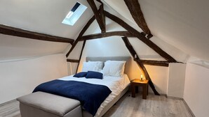 Een slaapkamer met een schuine kap, een bed, een nachtkastje met een lamp en een klein raam.