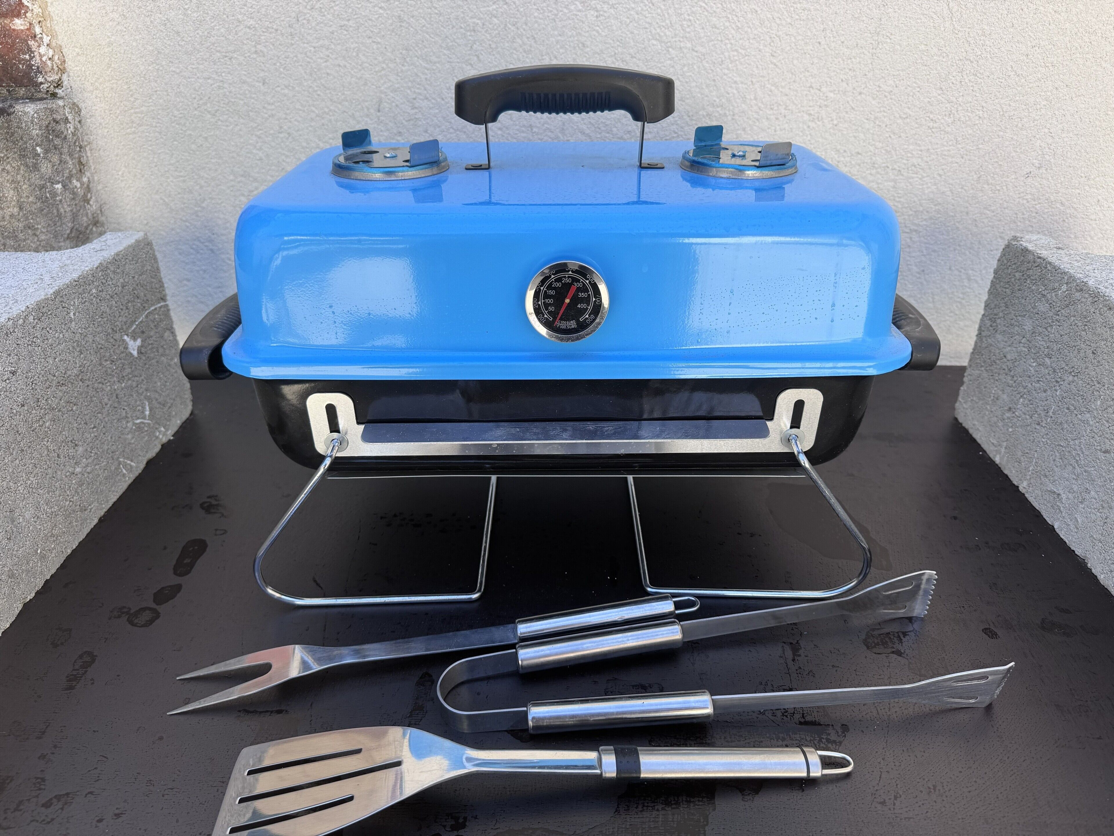 A blue portable grill with a digital thermometer, two metal spatulas, and a handle.