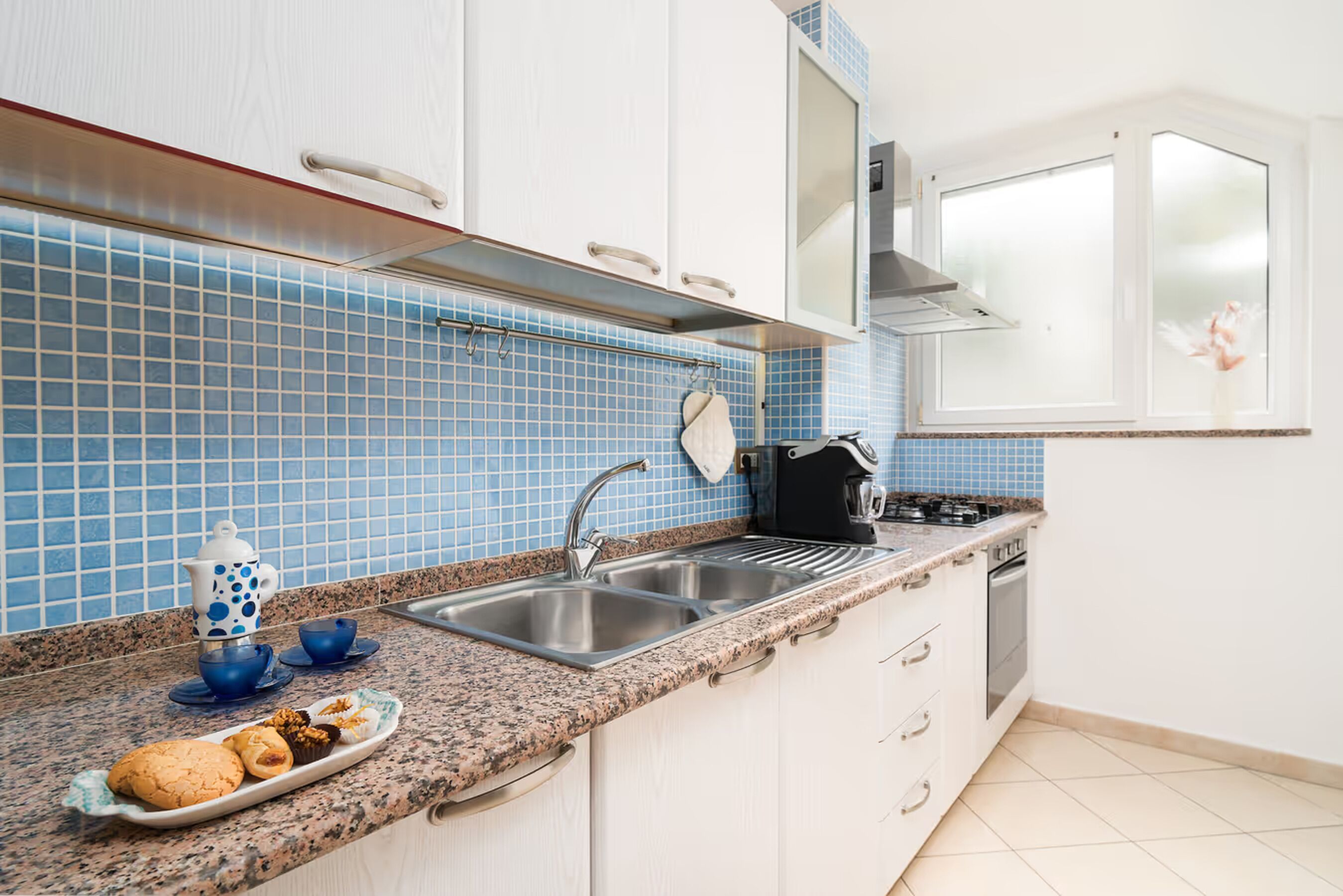 Una cocina con armarios blancos, salpicadero de azulejos azules, un lavamanos de acero inoxidable y una estufa.