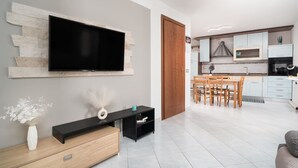 Ein modernes Wohnzimmer mit Flachbildfernseher, einer Holzkommode, einem Essbereich mit Tisch und Stühlen sowie einer Küche mit weißen Schränken und Geräten.