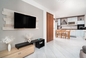 Una sala moderna con un televisor de pantalla plana, un mueble de madera, una zona de comedor con mesa y sillas, y una cocina con alacenas y electrodomésticos blancos.