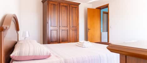 A bedroom with a bed, a wooden wardrobe, a bedside table, and a door leading to another room.