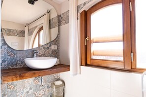 Ein Badezimmer mit weißem Waschbecken, einem Holzkosmetikschrank, einem Spiegel, gemusterten Fliesen, einem Fenster mit Blick und einer Tür.