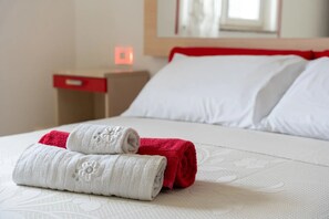 Een netjes opgemaakt bed met witte en rode handdoeken, een rode nachtkastje met een lade, en een witte muur met een raam.