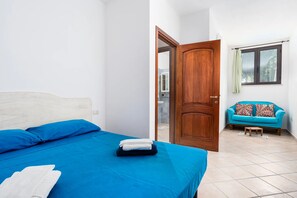 A bedroom with a bed, a wooden door, a window with a curtain, and a sofa in the adjacent room. - Su Nidu Virde - Four Bedroom House, Sleeps 9 (Orosei)