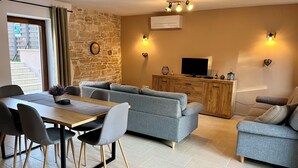 Ein Wohnzimmer mit Steinwand, einer Sofagarnitur, einem Esstisch mit Stühlen, einem Fernseher auf einem Holzschrank und einer Deckenleuchte.