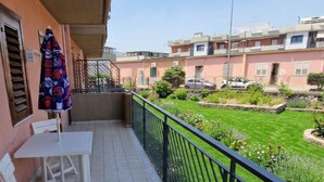 Un balcon avec une table et des chaises blanches, un parasol à motifs et une vue sur un jardin bien entretenu et des bâtiments en arrière-plan.
