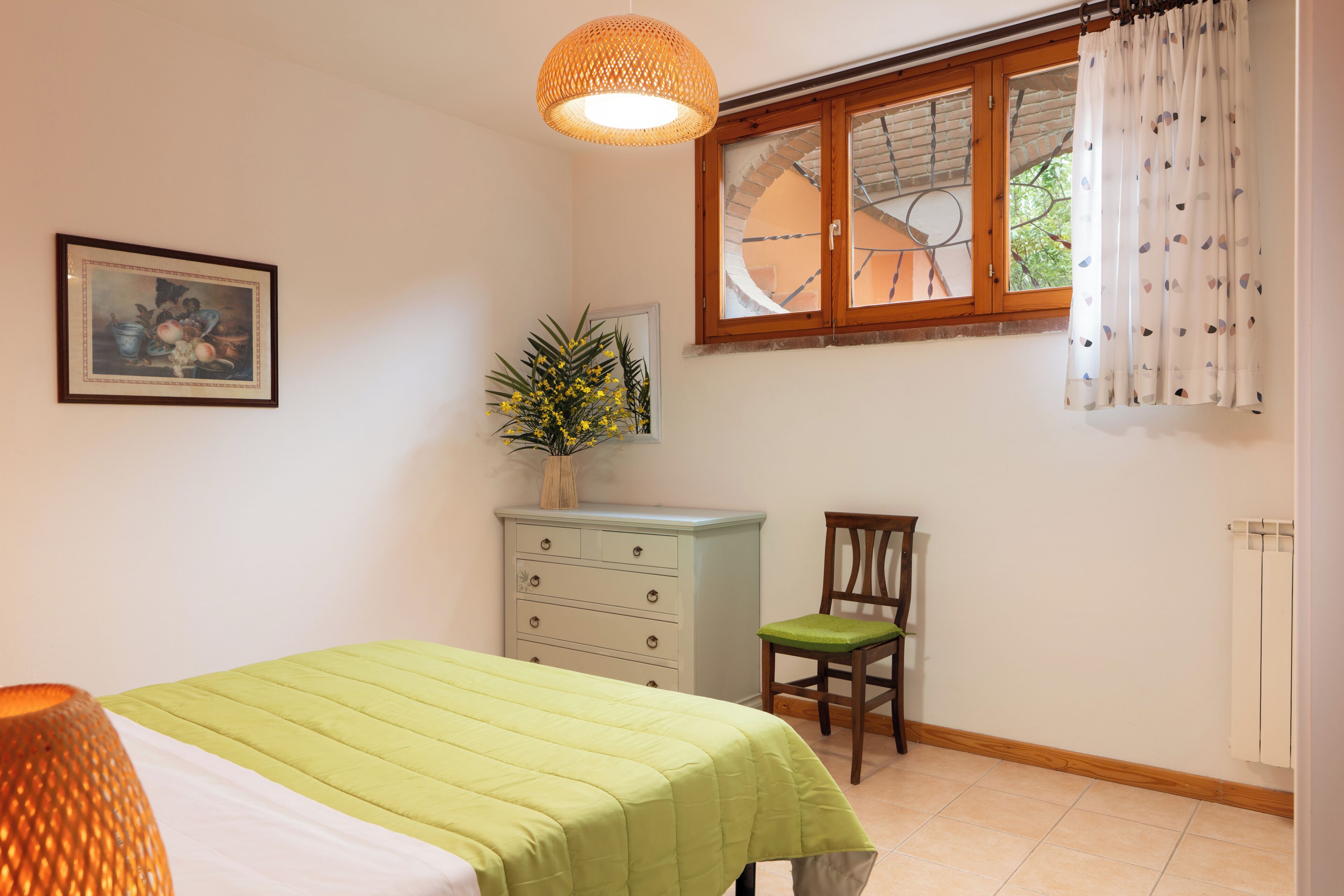 A bedroom with a bed, a dresser, a chair, a window with curtains, and a hanging light fixture.