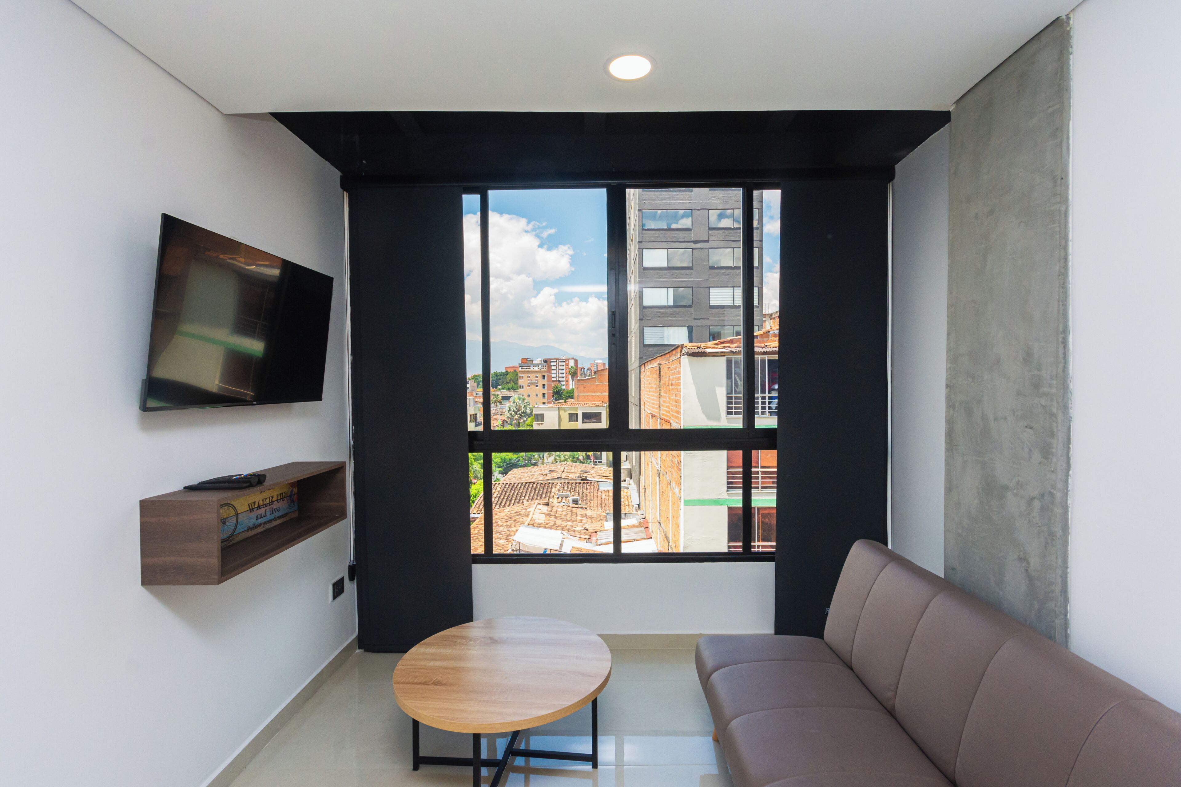 A modern living room with a sofa, a round table, a mounted TV, and a large window offering a city view.