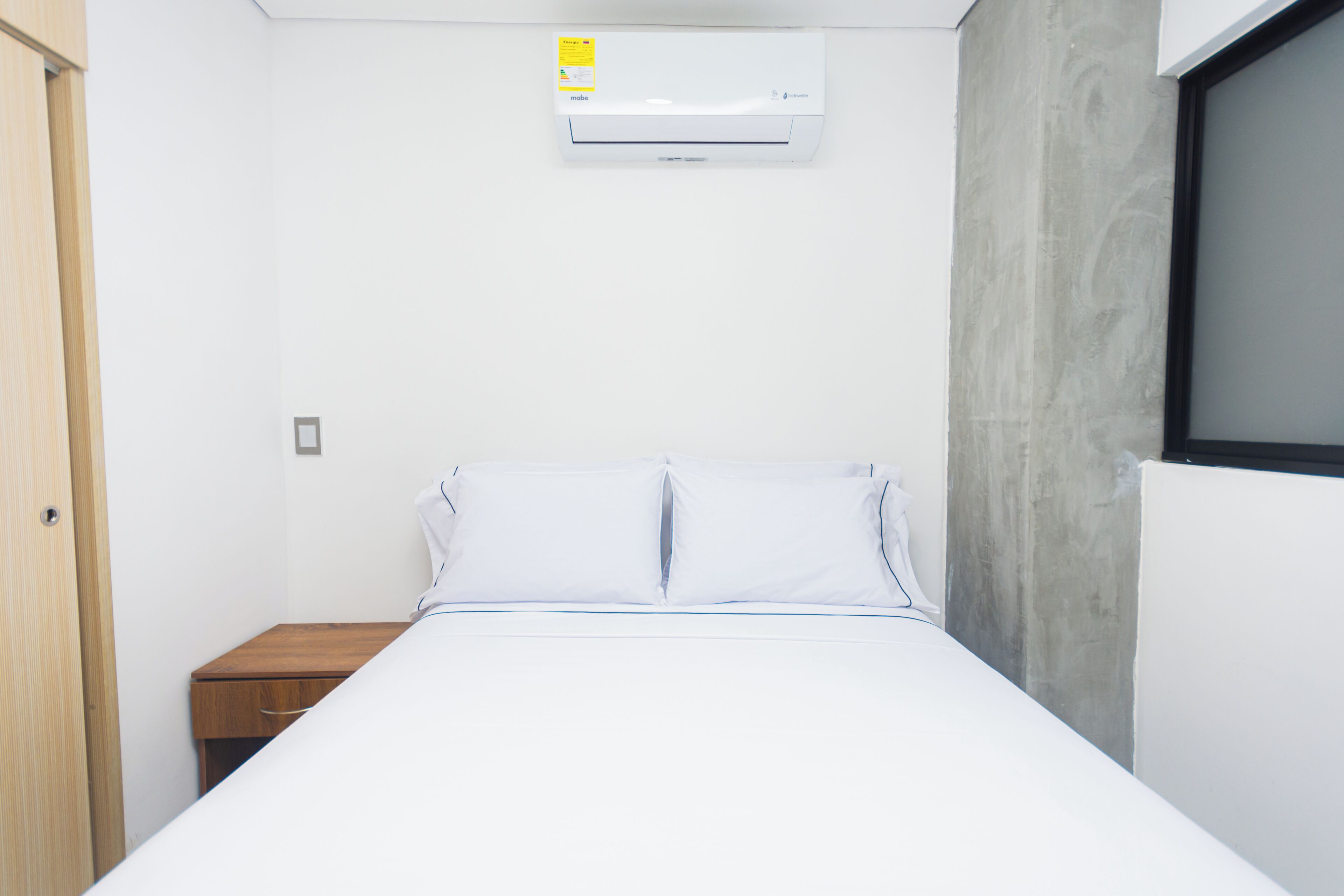 A bedroom with a bed, a wooden nightstand, a wall-mounted air conditioner, and a window.