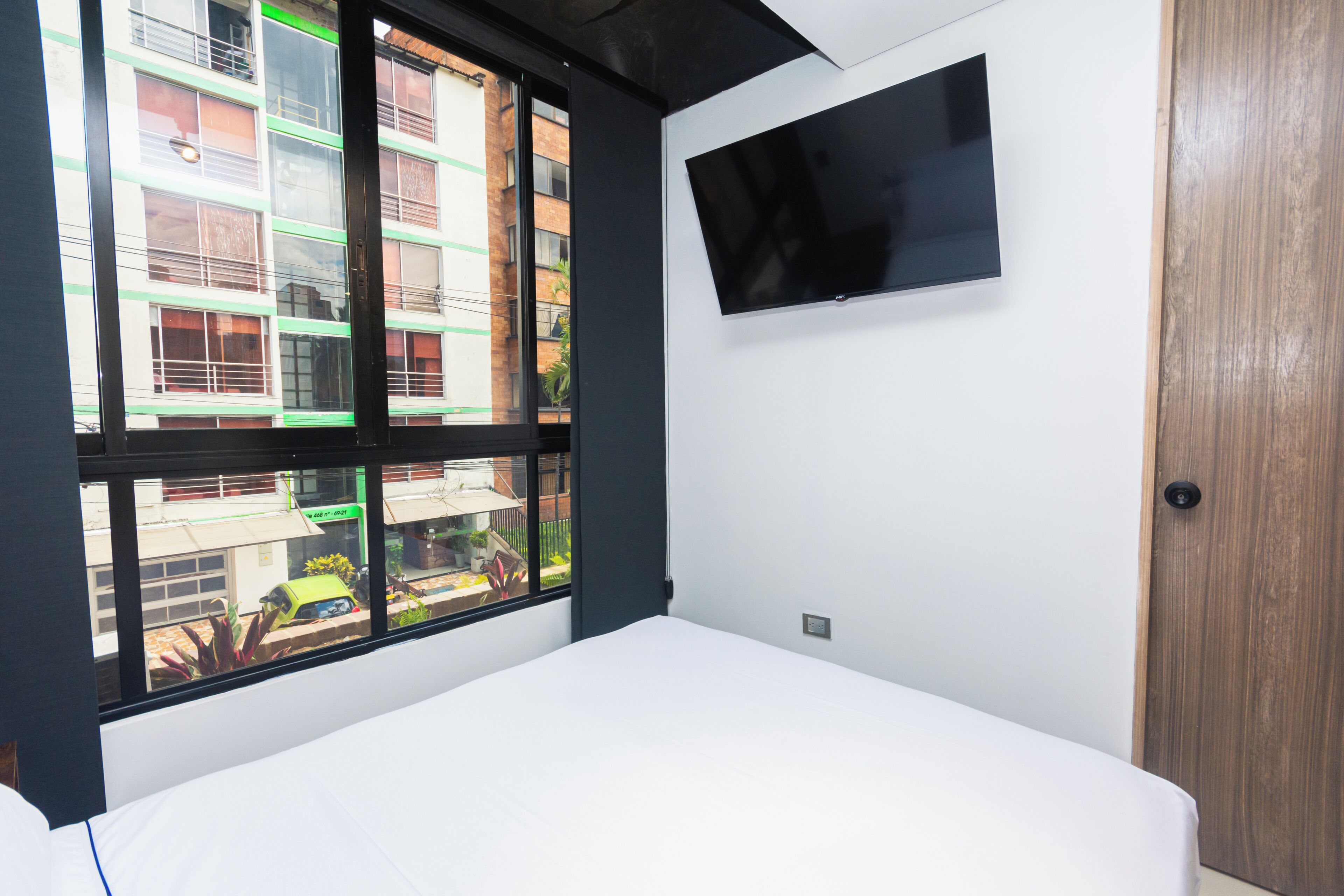 A room with a bed, a TV mounted on the wall, and a window with a view of buildings.
