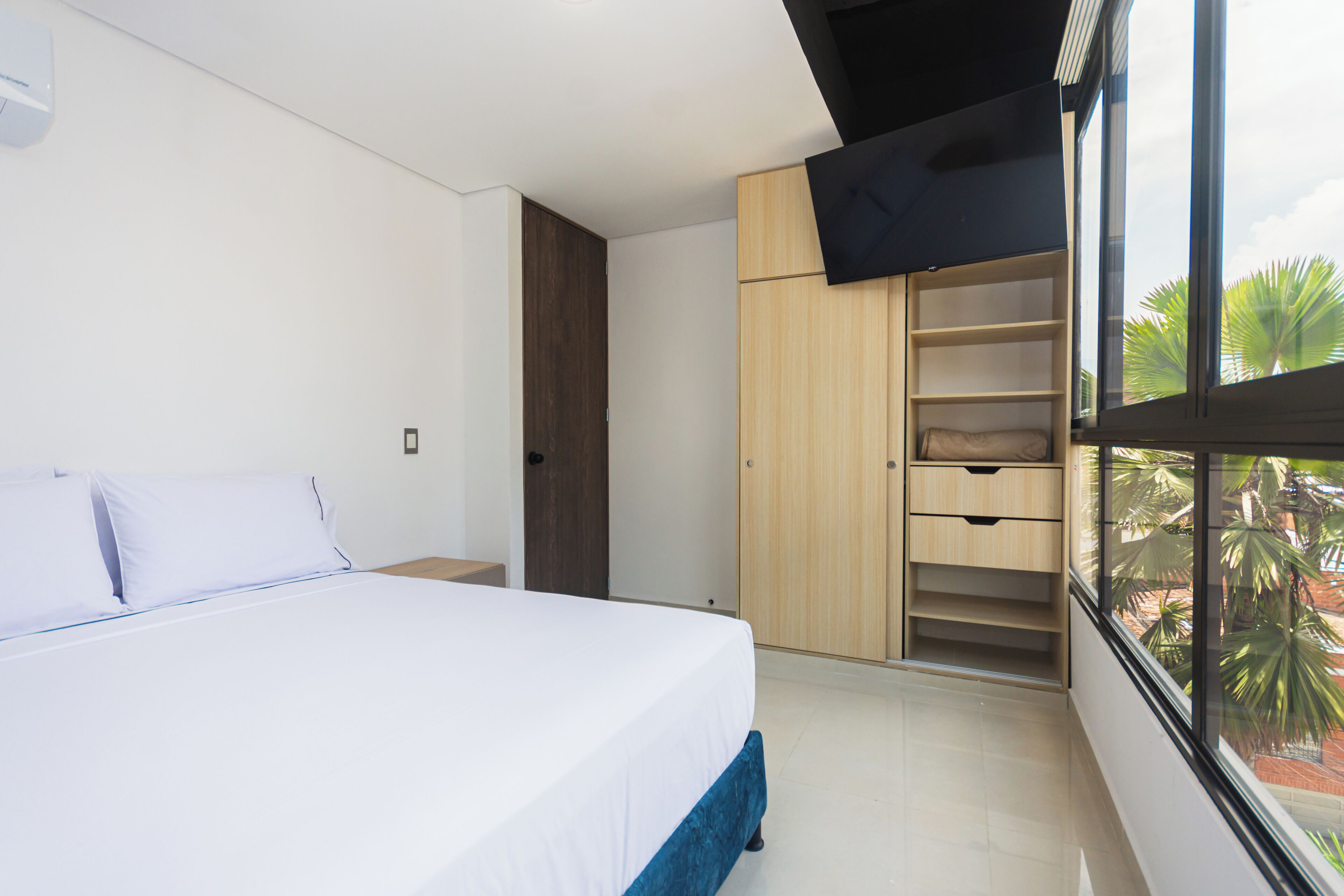 A bedroom with a bed, a wardrobe, and a window with a view of palm trees.