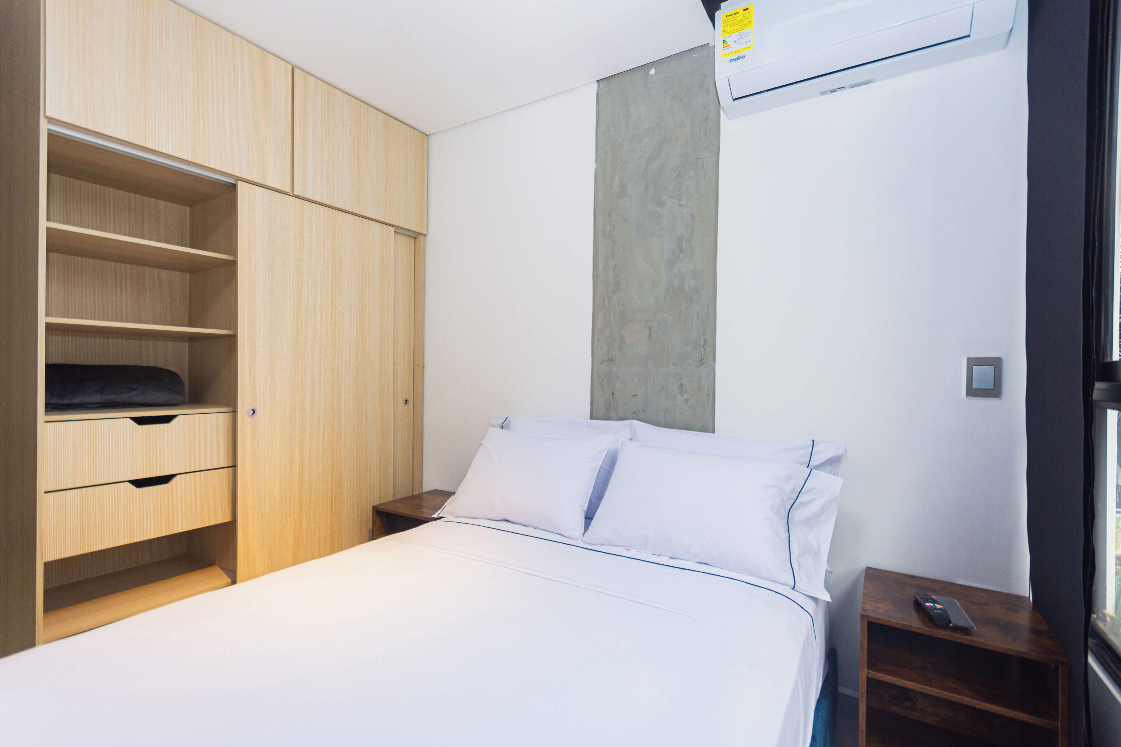 A bedroom with a bed, a wooden wardrobe, a nightstand, and a wall-mounted air conditioner.