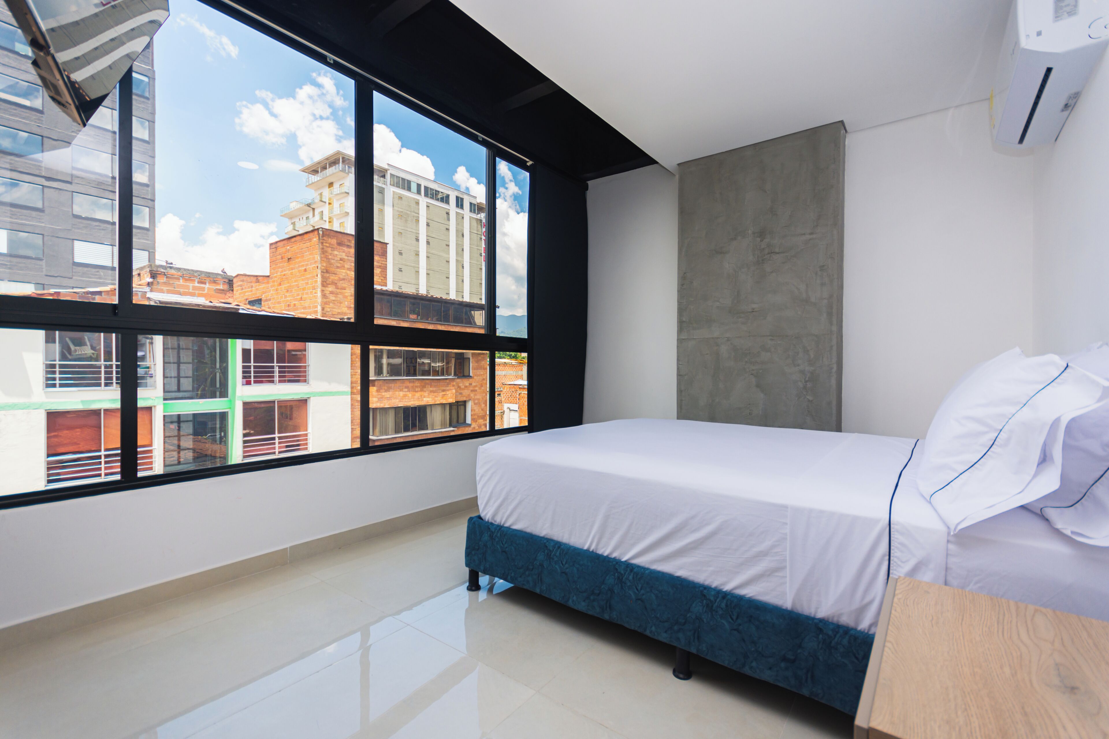A modern bedroom with a large window, a bed, a wooden nightstand, and a concrete accent wall.