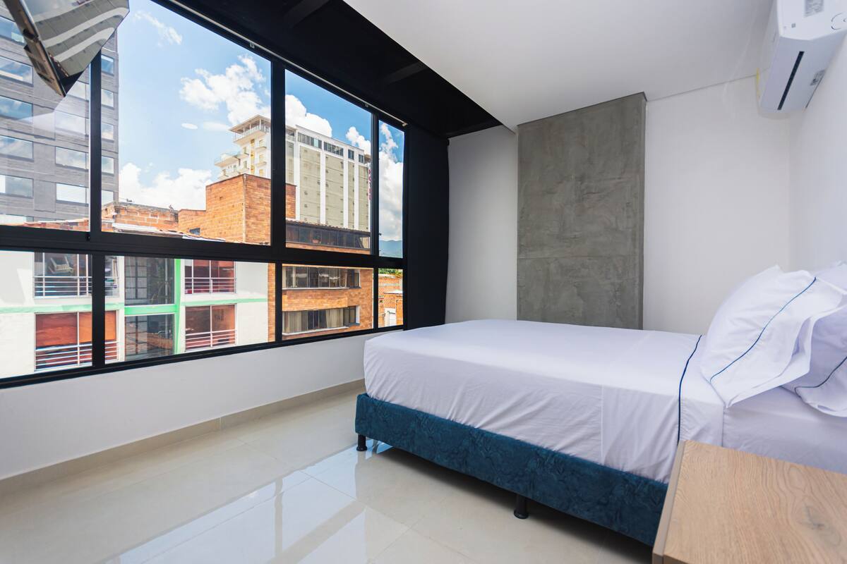 Un dormitorio moderno con un ventanal grande, una cama, una mesita de noche de madera y una pared de concreto decorativa.