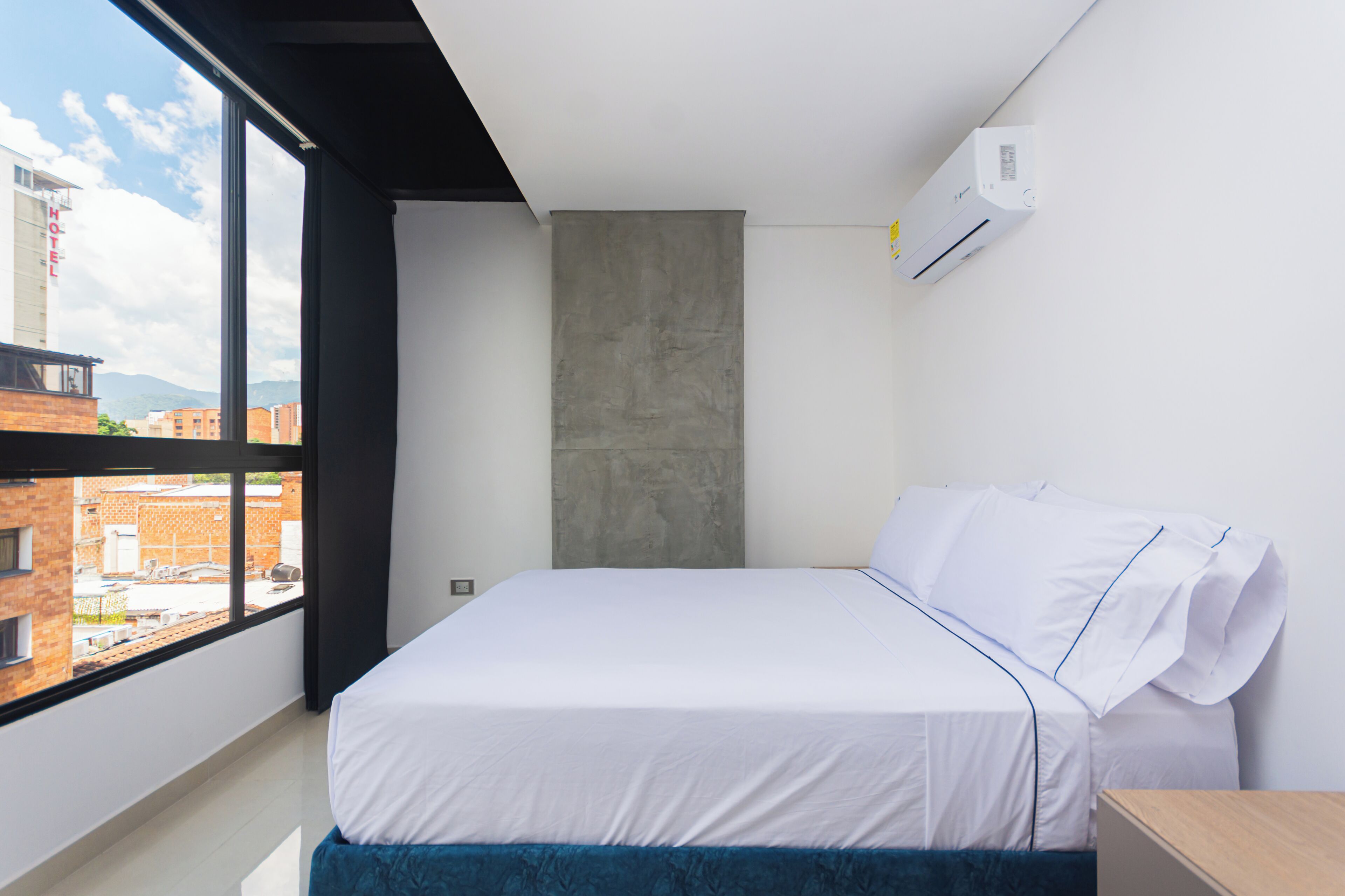 A modern hotel room with a large window, a bed with white linens, a concrete accent wall, and an air conditioning unit.