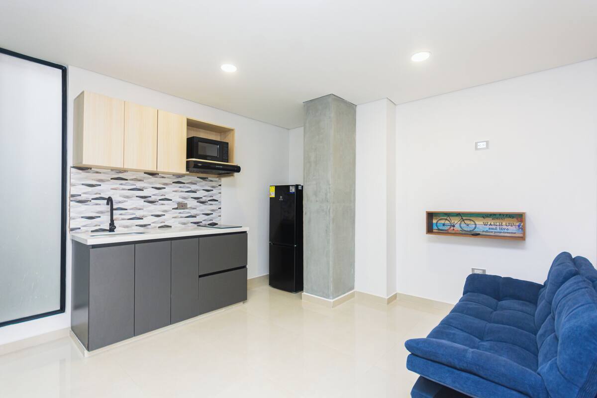 Una cocina moderna con armarios grises, un refrigerador negro y un microondas. En la zona de estar hay un sofá azul.