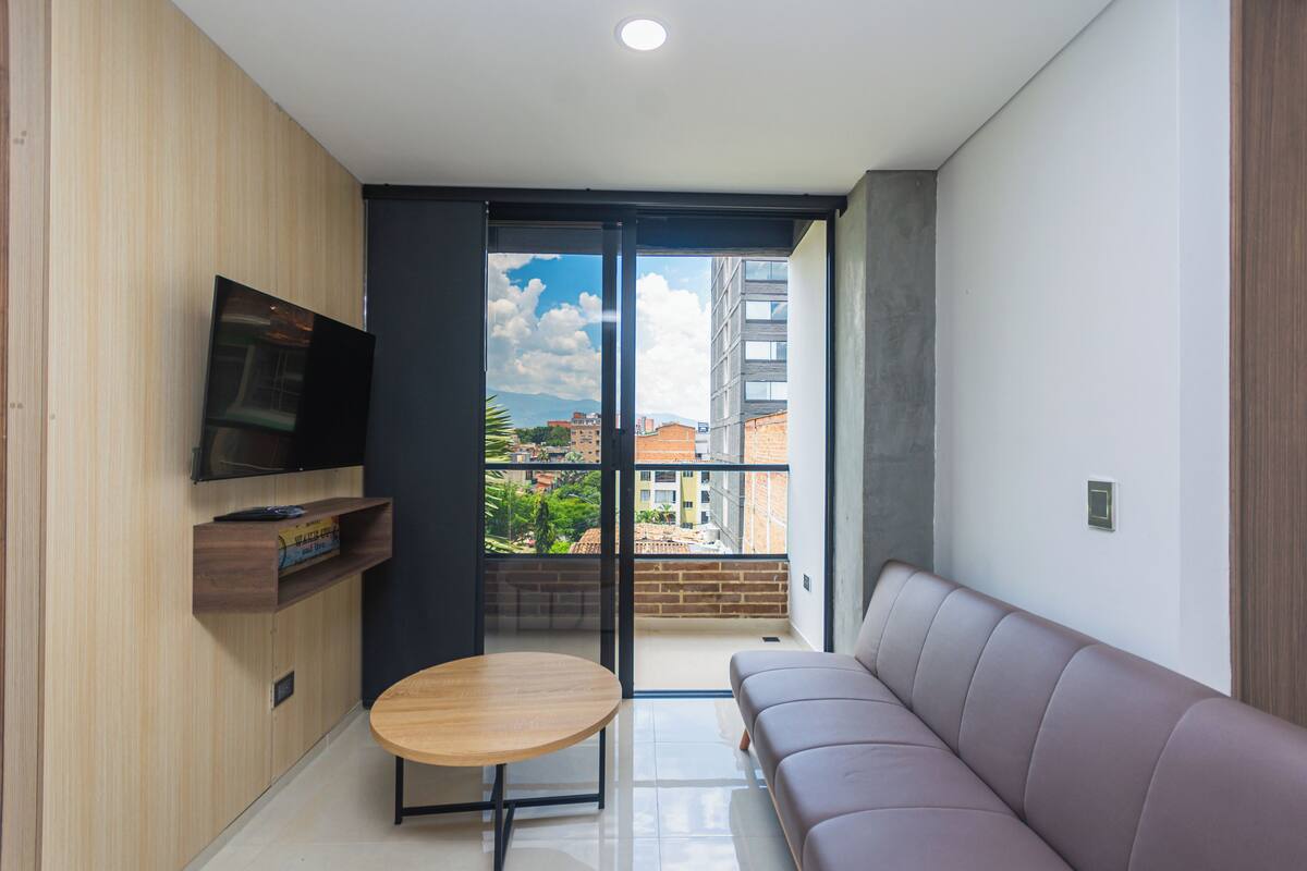 Una sala de estar moderna con un sofá, un televisor montado en la pared, una mesita de madera y un balcón con vistas a la ciudad.