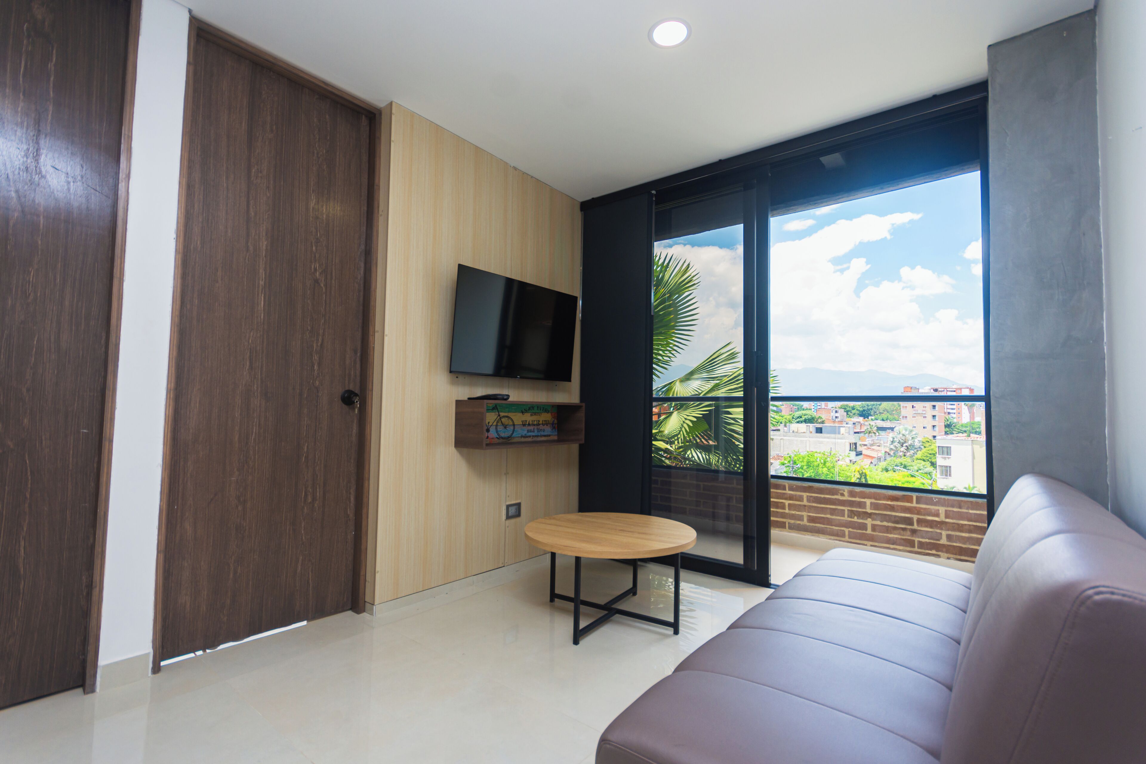A modern living room with a sofa, a small round table, a TV mounted on the wall, and a large window offering a city view.