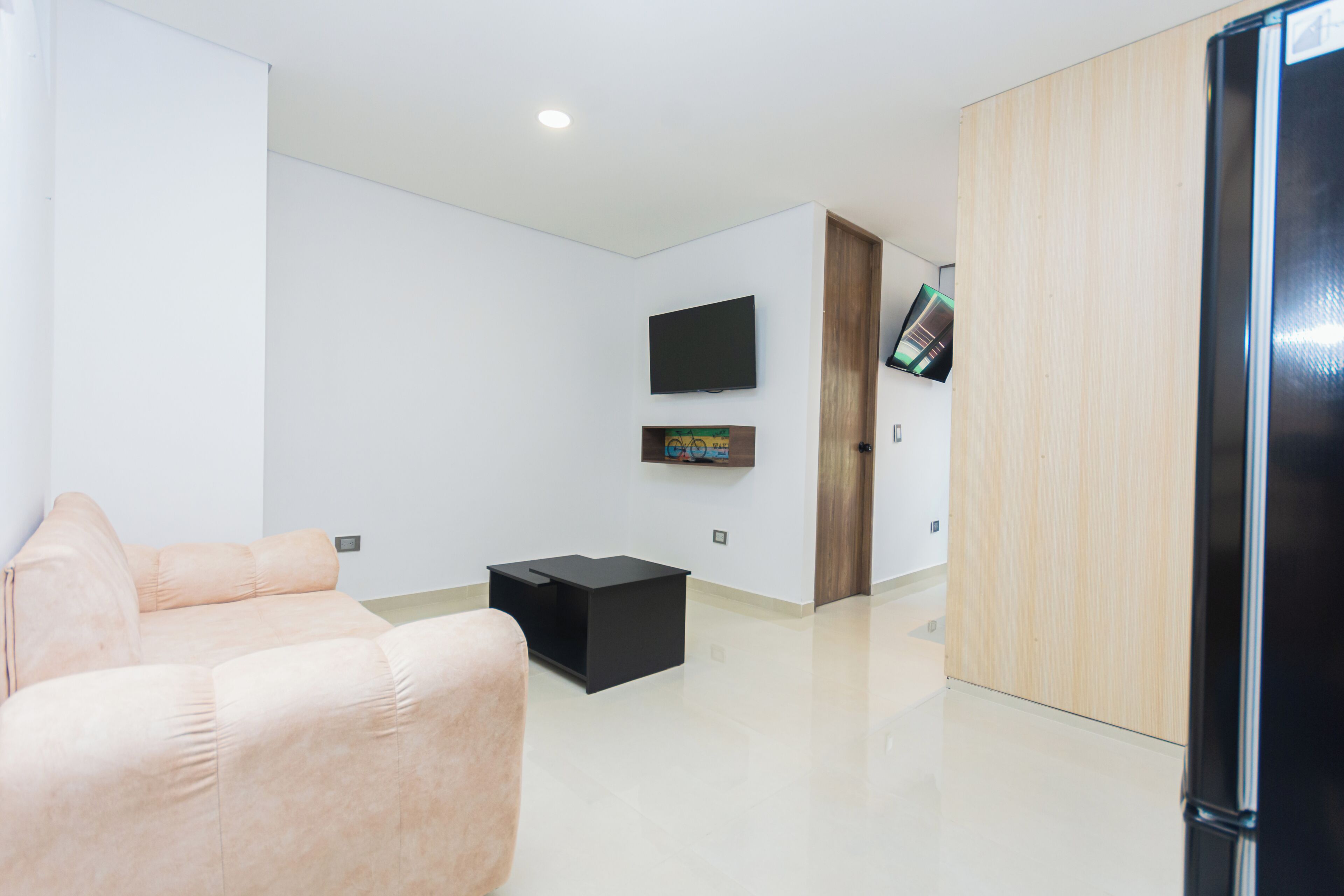 Een moderne woonkamer met een bank, een zwart voetenbankje, een flatscreen televisie aan de muur en een houten garderobe.