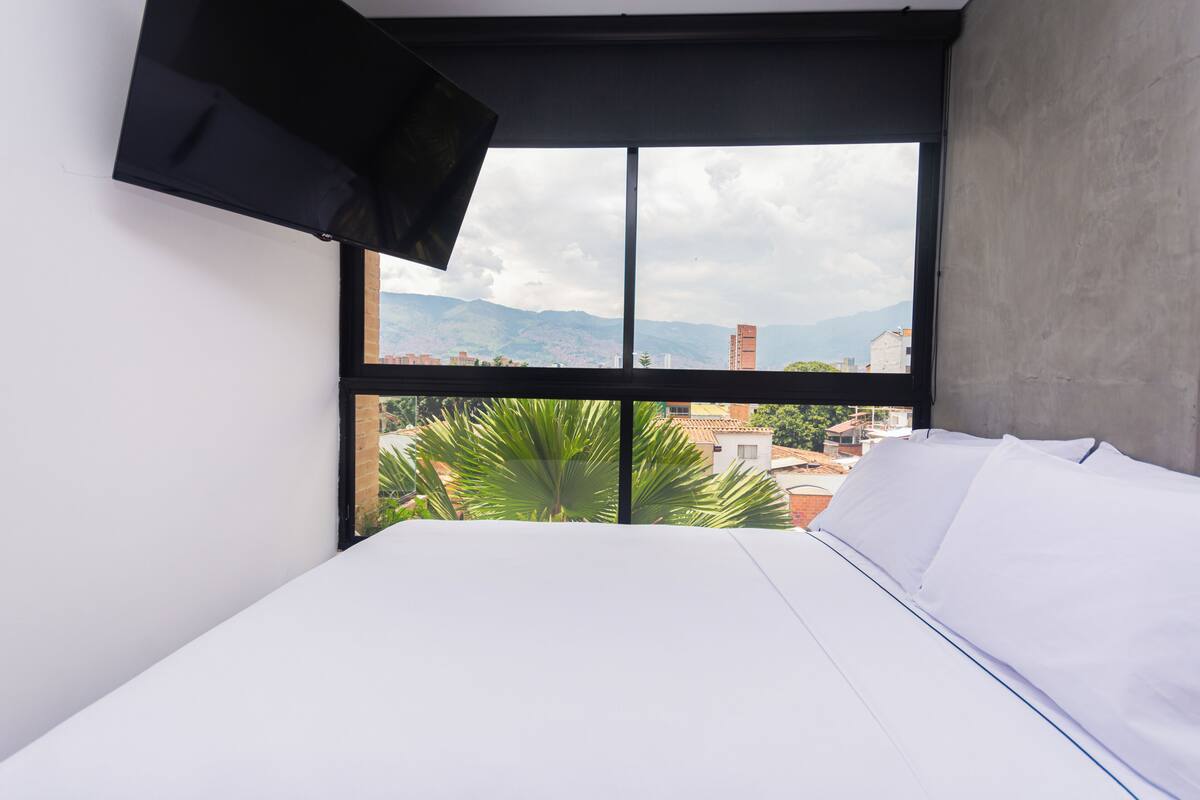 Un dormitorio con cama, ventana y vistas a edificios y montañas, y un televisor montado.
