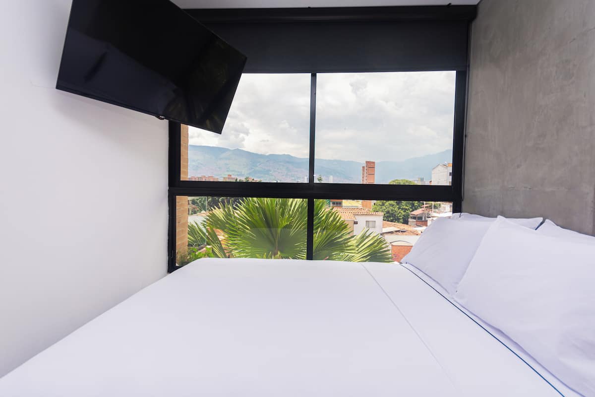 Un dormitorio con cama, ventana y vistas a edificios y montañas, y un televisor montado.