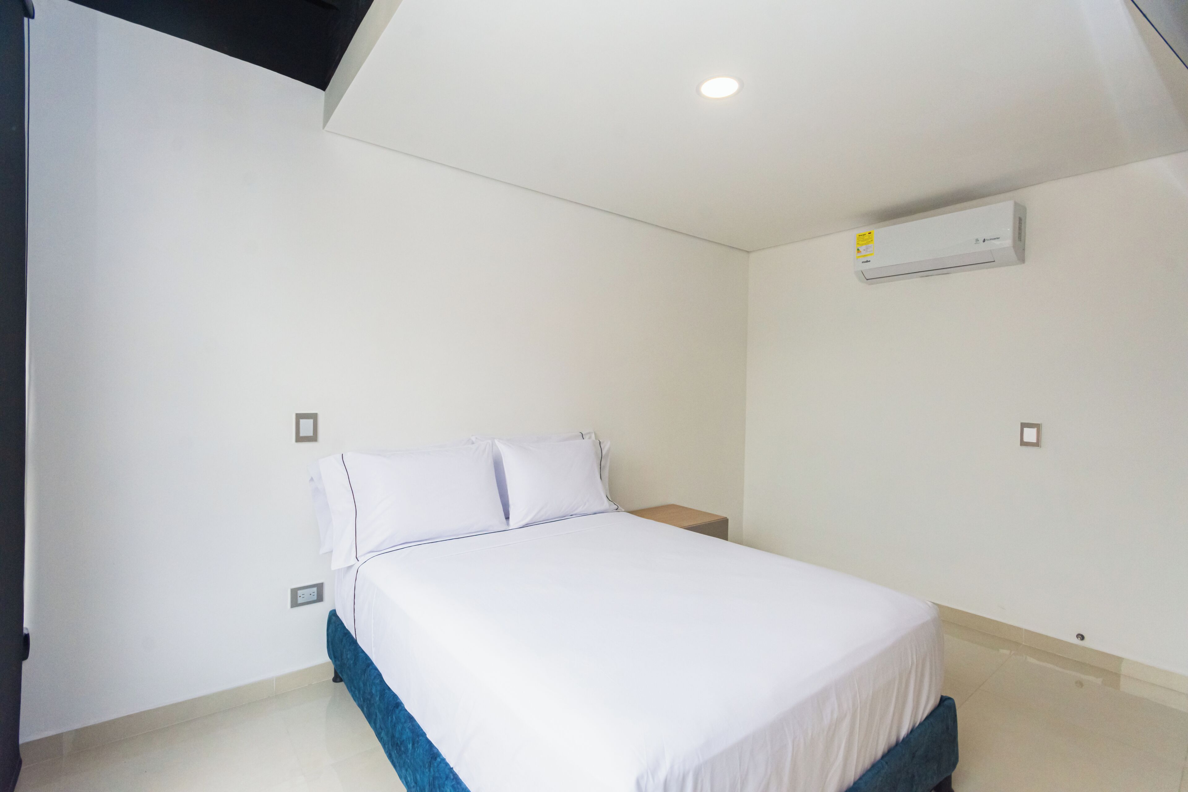 A bedroom with a bed, white walls, a ceiling light, and an air conditioning unit.