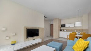 Een moderne woonkamer met een flatscreen televisie, een bank met gele kussens, een eethoek met een tafel en stoelen, en een keuken op de achtergrond.